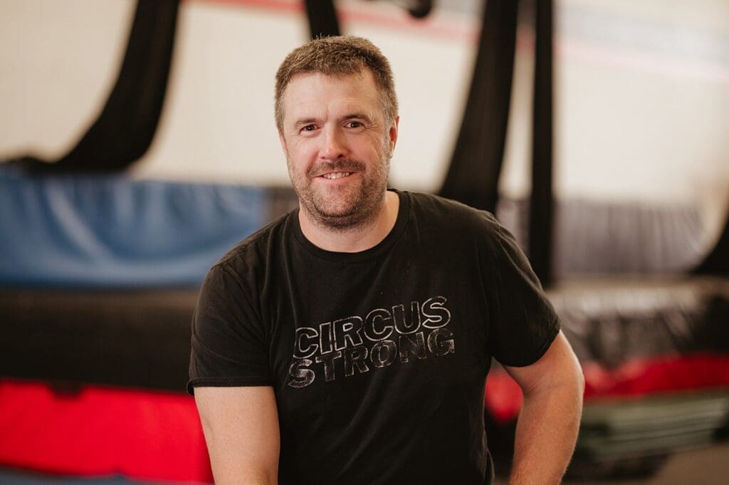 Headshot of a circus gym owner, smiling at the camera in a branded shirt with circus apparatus in the background.