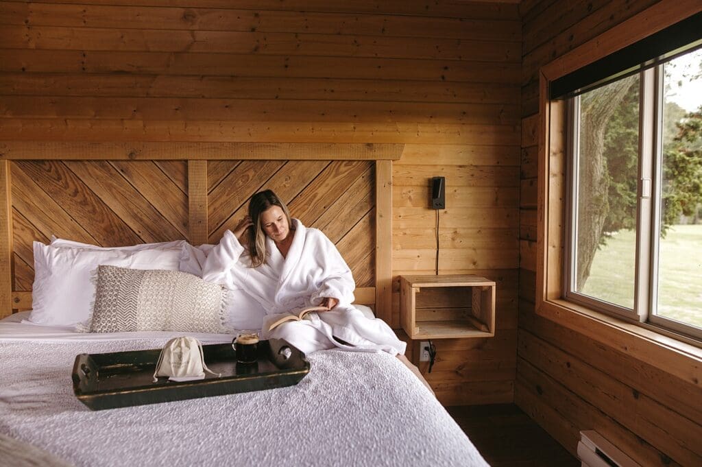 Woman wrapped in a cozy robe sitting on a beautiful bed in a cabin, reading and drinking coffee